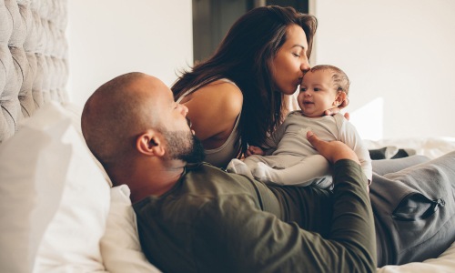 mother kisses her infant while father lies in bed and holds him
